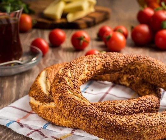 Simit ve Açma Kültürü Şehir Hayatında Değişmeyen Lezzetler Türkiye’de sokak lezzetleri denildiğinde akla gelen ilk tatlar arasında simit ve açma yer alır. Günün her saatinde ulaşılabilen bu iki hamur işi, yalnızca pratik bir atıştırmalık olmanın ötesinde, şehir yaşamının ritmini belirleyen önemli unsurlardan biri olarak görülür. Sabah işe yetişmeye çalışan bir memurun elinde, okul yolunda koşan bir öğrencinin çantasında ya da sahil kenarında çay eşliğinde tüketilen bu lezzetler, toplumsal hafızanın da güçlü bir parçasıdır. Konulu bir haber görseli.